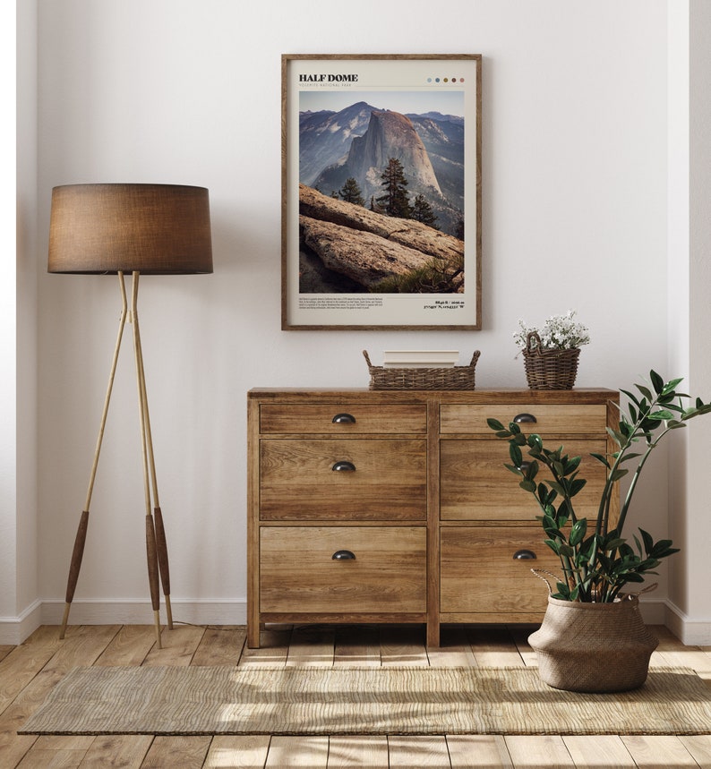May include: A framed print of Half Dome in Yosemite National Park hangs above a wooden dresser. The print features a landscape photograph with the text "HALF DOME" at the top. A floor lamp with a brown shade and a woven basket are also visible.