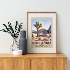 May include: A framed print featuring a Joshua Tree National Park landscape. The print displays a Joshua tree, rocks, and the text "Joshua Tree National Park". The frame is a light brown color, and the print is displayed on a wooden surface.