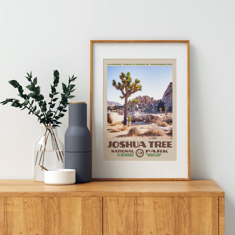 May include: A framed print featuring a Joshua Tree National Park landscape. The print displays a Joshua tree, rocks, and the text "Joshua Tree National Park". The frame is a light brown color, and the print is displayed on a wooden surface.