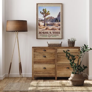 May include: A framed poster of Joshua Tree National Park hangs above a wooden dresser. The poster features a Joshua tree, desert landscape, and text. A floor lamp with a brown shade and a potted plant are also in the room.