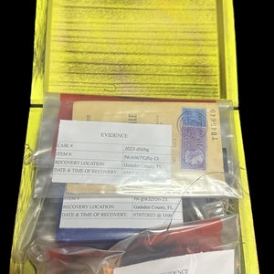 May include: A yellow wooden box with three clear plastic bags containing evidence. Each bag has a white label with the text "EVIDENCE" and details about the case, item number, recovery location, and date and time of recovery. The text on the labels includes: "2023-d56hg", "PA-niW7fQRq-23", "Gadsden County, FL", "07/07/2023 @ 1400", "PA-ghk3ZtDn-23", "07/07/2023 @ 1100", "PA-QaGNbdX4-23", "07/07/2023 @ 1100".
