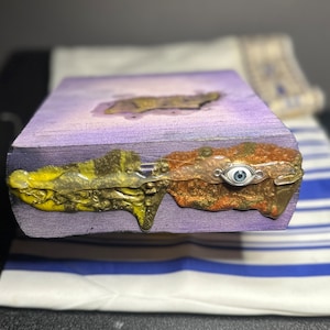 May include: A purple wooden box with a gold and yellow resin design and a blue evil eye charm. The box is sitting on a white and blue striped fabric.