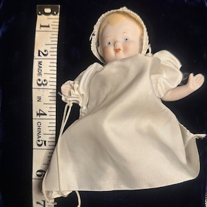 May include: A porcelain baby doll wearing a white satin dress with a lace trim. The doll is approximately 4 inches tall and is lying on a blue velvet surface.