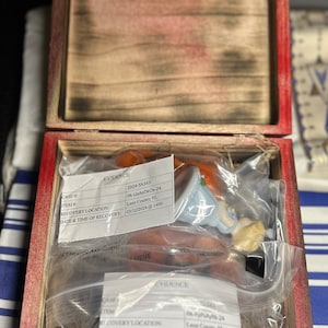 May include: Two small porcelain figurines of people in a blue dress and a white dress, packaged in a clear plastic bag with a white label that reads "EVIDENCE 2024-5h343 PA-UeAaDkOx-24 Leon County, FL 03/12/2024 @ 1400". The bag is inside a wooden box with a red lid.