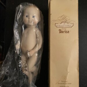 May include: A porcelain baby doll with blue eyes and a light pink complexion. The doll is packaged in a clear plastic bag and a white box with the text "The Collectors' Choice from Darice" printed on it.