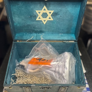 May include: A teal blue wooden treasure chest with brass accents and a gold star of David on the lid. The chest is open and contains a white porcelain doll wrapped in plastic.