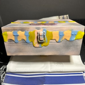 May include: A wooden box with a latch, painted in shades of blue, yellow, and pink. The box has a distressed wood finish and is decorated with a colorful abstract design.