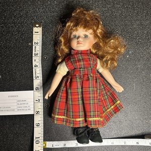 May include: A porcelain doll with red hair, green eyes, and a red and black plaid dress. The doll is wearing black shoes and is approximately 7 inches tall.