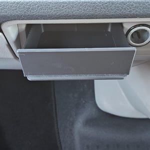 May include: A black, rectangular car console tray, pulled open, is positioned below the dashboard. The tray is made of a matte black material and is located near a power outlet and air vent controls.