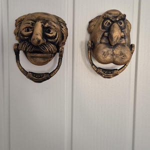 May include: Two antique brass door knockers, each depicting a stylized face with a handle below. One has a mustache, the other a large nose. The knockers are mounted on a white door.
