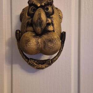 May include: A gold-coloured door knocker featuring a sculpted face with a large nose, prominent eyebrows, and a circular swinging ring at the bottom. The surface has a textured, layered appearance with dark shading in the recessed areas.