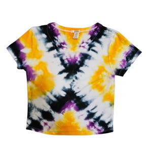 May include: A tie-dye crop top in a ribbed knit fabric. The shirt features a V-neck and short sleeves. The tie-dye pattern includes yellow, purple, black, and white colors. The shirt is a casual style.
