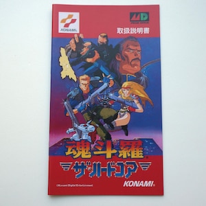 Pode incluir: Um manual de instruções de videogame retangular com fundo vermelho e azul. A capa apresenta personagens ilustrados em poses de ação, o logotipo da Konami, o logotipo MD e texto em japonês, incluindo o título 魂斗羅 ザ・ハードコア.