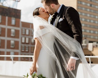 Può includere: Una sposa e uno sposo si baciano davanti a un edificio con un cartello che dice "ELERS". La sposa indossa un abito bianco con un velo e tiene un bouquet di rose bianche. Lo sposo indossa uno smoking nero con un papillon nero.