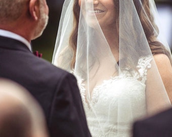 May include: A bride wearing a white wedding dress with a veil covering her face smiles as she looks at the person in front of her.