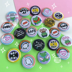 May include: A collection of colorful round buttons with various messages and illustrations related to LGBTQ+ pride. The buttons feature phrases like "Queer AF", "Not a Girl", "You Are Safe", "We Have Always Existed", and "Pride Started as a Protest". Some buttons also include illustrations of frogs, hearts, and rainbows.