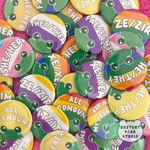 May include: A collection of colorful pronoun buttons with frog illustrations. The buttons feature phrases like "They/Them," "She/Her," and "All Pronouns." The buttons are in various colors, including rainbow, purple, yellow, and green.