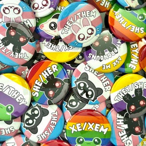 May include: A collection of round pin-back buttons featuring various pride flag color backgrounds and cartoon animal illustrations. Each button displays specific pronouns such as they/them, she/her, xe/xem, or all pronouns in bold white text, alongside cute characters like frogs, rabbits, and moths.