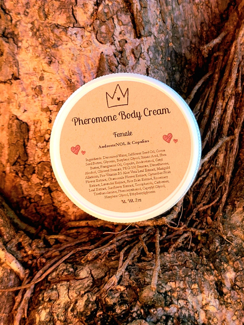 May include: A brown jar with a white lid and a label that reads "Pheromone Body Cream Female AndrosteNOL & Copulins". The jar is sitting on a tree trunk.