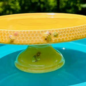 May include: A yellow ceramic cake stand with a honeycomb design and bee accents. The stand is sitting on a blue surface.