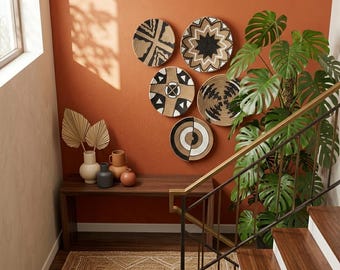 May include: A home decor scene featuring six woven wall baskets with geometric patterns in black and natural tones. A wooden bench holds vases and dried leaves. A large potted plant and a staircase with a brass railing are also visible.