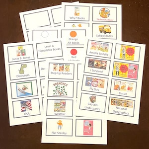 May include: A collection of white cards featuring book covers and titles, including "Junie B. Jones," "Dinosaurs," and "Flat Stanley." The cards are arranged on a brown surface, with some labeled "Level A Decodable Books."