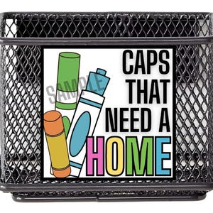 May include: A black wire mesh desk organizer with a rectangular shape. The organizer features a colorful graphic with the text "CAPS THAT NEED A HOME" and illustrations of markers and caps.
