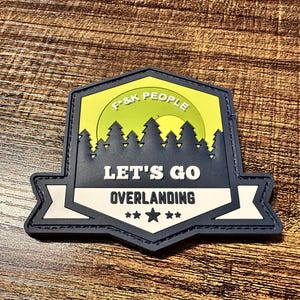 May include: A navy blue and white patch with the text "LET'S GO OVERLANDING" and the words "F&K PEOPLE" in a yellow and green background. The patch features a forest silhouette and three stars.