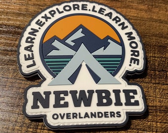 May include: White patch with black text that reads "NEWBIE OVERLANDERS". The patch has a graphic of a tent in front of mountains with a sunset behind them. The text "LEARN. EXPLORE. LEARN MORE." is around the graphic.