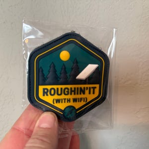 May include: A patch with a dark blue border and a yellow outline. The patch has a teal background with a yellow sun, green trees, and a white Wi-Fi symbol. Text on the patch reads "ROUGHIN'IT (WITH WIFI)".