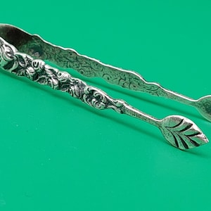 Puede incluir: Pinzas de azúcar de plata con un diseño floral ornamentado. Las pinzas están marcadas con el número "800" en el mango.