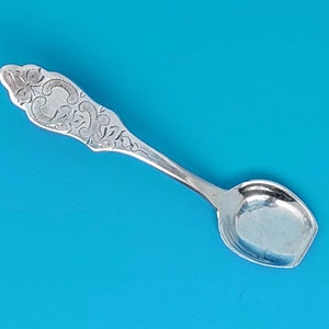 Può includere: Un cucchiaino da sale d'argento con un manico decorato e inciso e una ciotola arrotondata. Il cucchiaino è su uno sfondo blu.