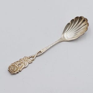 May include: A silver sugar spoon with a rose design on the handle. The spoon has a scalloped bowl.