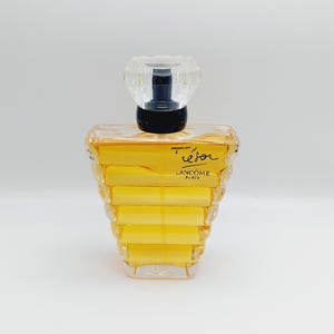 Könnte beinhalten: Ein klarer Glasparfümflakon mit einem facettierten, kristallartigen Verschluss. Die Flasche enthält eine gelbe Flüssigkeit und das Wort "Trésor" ist auf der Vorderseite zusammen mit "Lancôme Paris" geschrieben. Die Flasche hat ein einzigartiges, gestuftes Design.