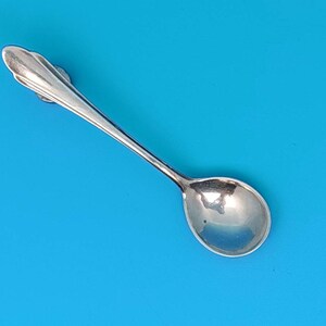 May include: A small, silver spoon with a rounded bowl and a decorative handle. The spoon is set against a bright blue background, highlighting its polished surface. The spoon's design suggests it is for serving or measuring small quantities.