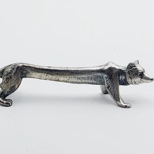 Peut inclure: Un repose-baguettes en métal argenté en forme de chien allongé. La figurine présente des textures de fourrure détaillées sur la tête et les pattes, avec un long corps lisse et arqué reliant les pattes avant et arrière dans une pose étirée.