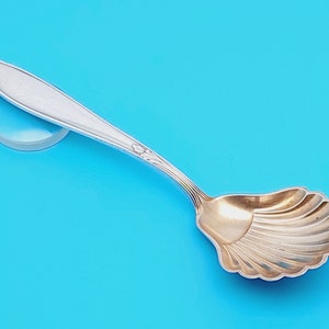 Pode incluir: Uma colher de açúcar de prata com uma tigela dourada e festonada. A colher tem um design simples e elegante.