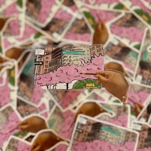 May include: A collection of small, rectangular art prints featuring a cityscape with pink cherry blossom trees. Each print is being painted with a brush, with hands holding the brush. The prints are layered on top of each other.
