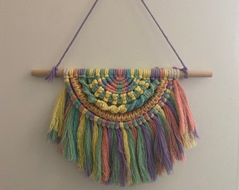 Puede incluir: Un tapiz de macramé de pared colorido con un arcoíris de colores, incluyendo amarillo, verde, rosa y morado. El tapiz presenta un diseño de medio círculo con borlas en la parte inferior.