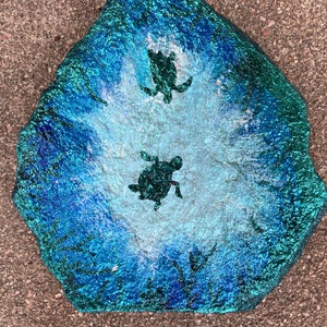 May include: A painted rock with a blue and green ocean scene. Two black silhouettes of sea turtles are swimming in the water.