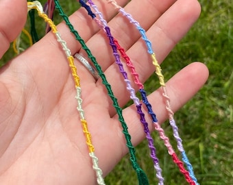 May include: A close-up of a hand holding seven colorful friendship bracelets. The bracelets are made of thin, braided string in shades of green, yellow, pink, blue, purple, and white.