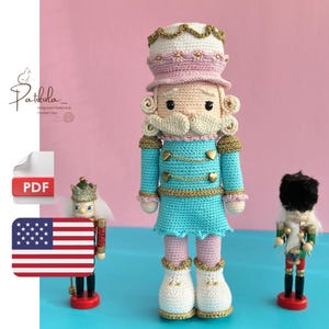 Puede incluir: Muñeca cascanueces de ganchillo con uniforme azul turquesa con ribetes dorados, detalles rosas y sombrero blanco y rosa. La muñeca tiene bigote blanco y está junto a dos figuras de cascanueces más pequeñas. El fondo es rosa liso.