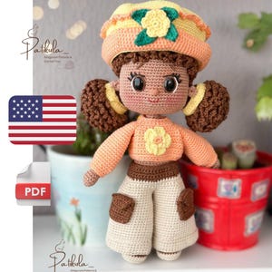 Può includere: Bambola all'uncinetto fatta a mano con capelli castani raccolti in due chignon, indossa un maglione color pesca, pantaloni color crema e un cappello abbinato. La bambola ha un fiore sul maglione e sul cappello. È un modello amigurumi.