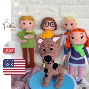 May include: A collection of crocheted amigurumi figures depicting cartoon characters. The figures include a brown dog with black spots, and four human characters. The handmade figures have detailed features and bright colours. The background is blurred.