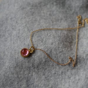 May include: A delicate gold chain necklace featuring a single round, translucent pink gemstone pendant set in a polished gold bezel. The fine metal chain connects to the circular setting, which holds the smooth, cabochon-style pink stone securely.
