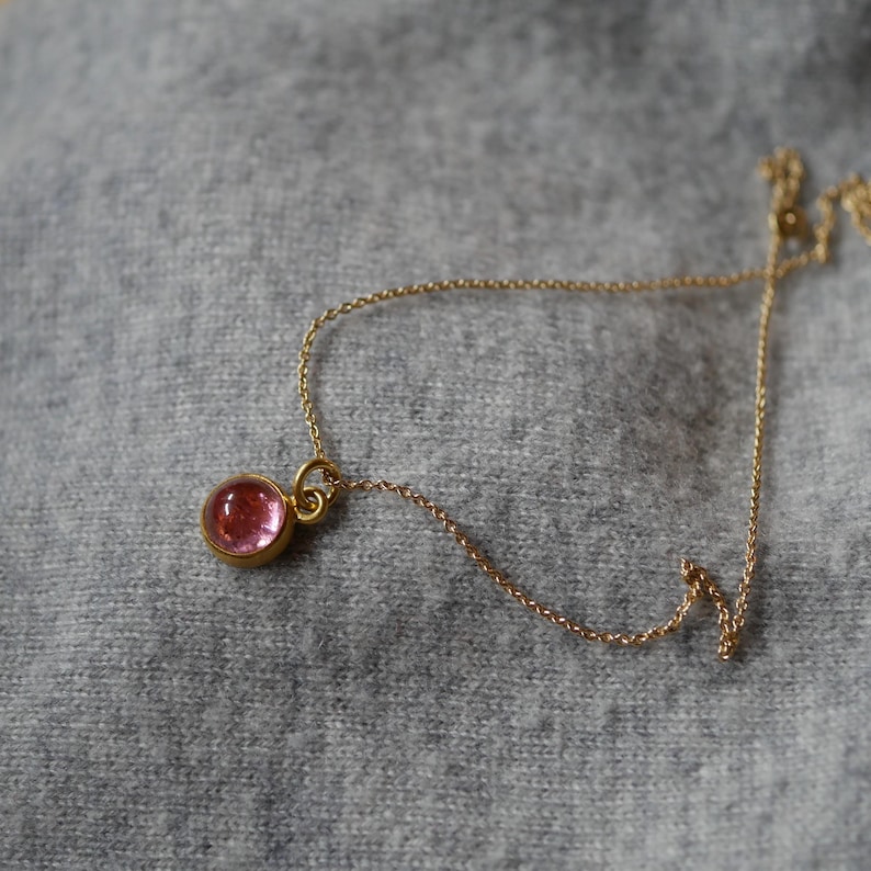 May include: A delicate gold chain necklace featuring a single round, translucent pink gemstone pendant set in a polished gold bezel. The fine metal chain connects to the circular setting, which holds the smooth, cabochon-style pink stone securely.