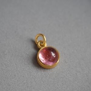 May include: A small, round pendant featuring a translucent pink gemstone set in a smooth, polished gold-toned metal bezel. The pendant includes a circular gold-toned jump ring attached to the top, designed for easy connection to a chain or cord.