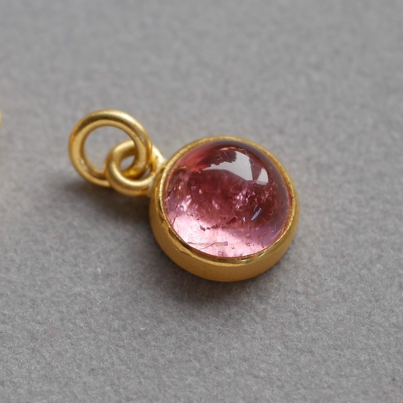 May include: A small, round pendant featuring a polished, translucent pink gemstone set in a smooth, gold-toned metal bezel. The pendant includes a circular gold-toned jump ring attached to the top, designed for easy connection to a necklace chain.