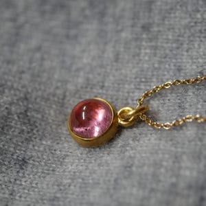 May include: A gold pendant necklace featuring a round, cabochon-cut pink gemstone set in a smooth gold bezel. The pendant is attached to a delicate gold chain, showcasing a polished finish and a vibrant, translucent pink hue on the stone.