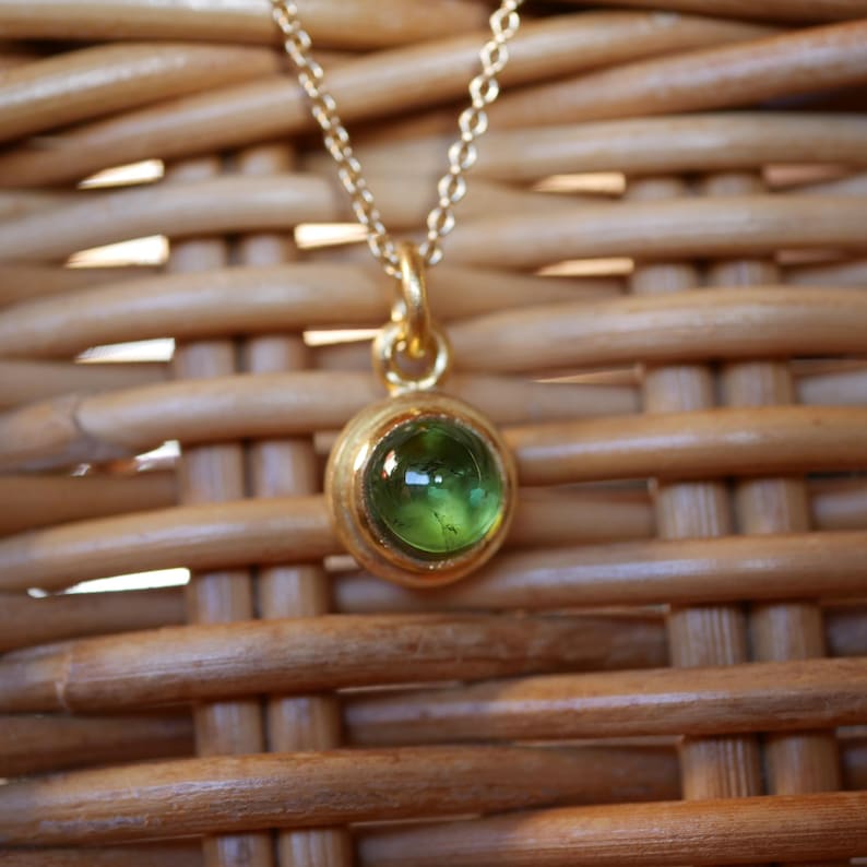 Puede incluir: Un collar de oro con un peque&ntilde;o colgante de piedra verde redonda en engaste de bisel. El colgante cuelga de una delicada cadena de oro, mostrando un acabado liso y pulido en el engaste met&aacute;lico que sostiene la piedra verde transl&uacute;cida.